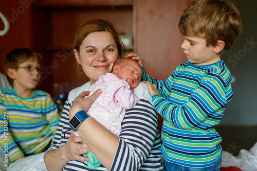 Mother holding newborn baby girl in hospital bed, with two sons sitting beside her. Real life, real people moment of family love, siblings bonding, and joy of welcoming a new child.