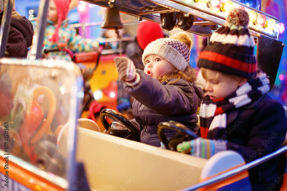 Naklejka premium Adorable little boy and girl, siblings on a carousel at Christmas funfair or market, outdoors. Happy children, friends having fun. Selective focus on one child. Holiday, children, lifestyle concept.