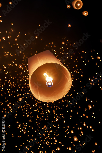 hot air sky lantern (Yee Peng) rising high into the dark night sky, surrounded by hundreds of floating lights. Focus is on the warm glow of the fire.