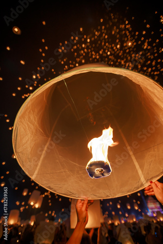People launching a hot air sky lantern (Yee Peng) into the night sky during a mass release festival in Thailand. The frame captures the fire and many floating lights.