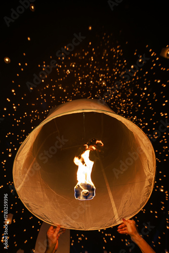 People launching a hot air sky lantern (Yee Peng) into the night sky during a mass release festival in Thailand. The frame captures the fire and many floating lights.