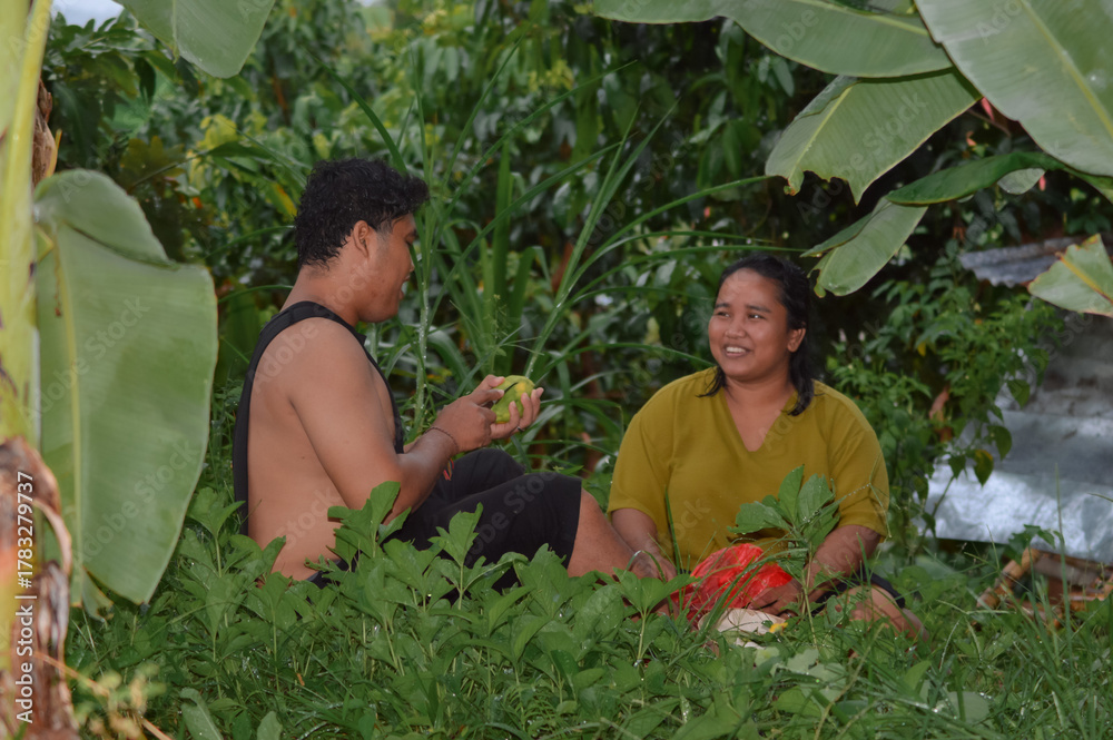 Fototapeta premium Husband and wife sit together on damp grass, talking softly while he peels a mango, sharing comfort and laughter after their long workday in the orchard.