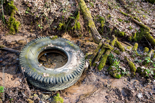 Grungy, discarded car tire half-submerged in a muddy forest creek, symbolizing environmental pollution and neglect. A bleak contrast of human waste amidst natural decay