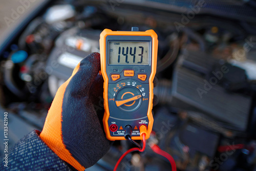 Man testing alternator charge and battery voltage with handheld diagnostic tool while car engine idles seasonal check for automotive electrical system repair maintenance and safe winter driving. 14v
