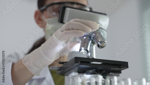 Female scientist wearing white protective gear analyzing test samples under microscope in contemporary research environment, close up of white gloved hand. Medicine and science concept