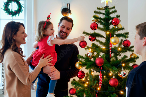 Caucasian woman holding young Caucasian girl while Caucasian man and teenage boy decorating Christmas tree together, smiling and interacting in festive home setting