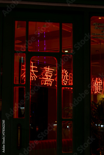 Red neon Chinese characters glow in a Bangkok Chinatown shop window at night. Reflections capture the bustling energy of the night market.