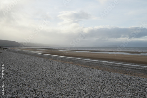 Vast Pebble And Sandy Beach With Calm Sea And Hazy Cloudy Sky Stretching Towards Distant Misty Hills In Towyn, North Wales, Uk.