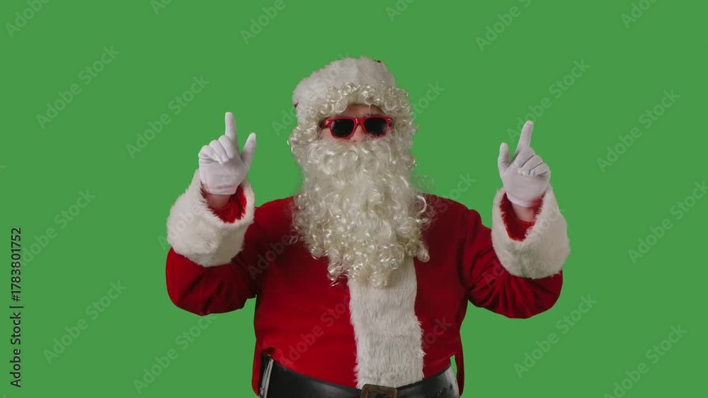 Santa Claus with a white curly beard, wearing a red suit and sunglasses, points his fingers upward on a green background. Perfect for Christmas themed compositions, advertising, and holiday layouts.
