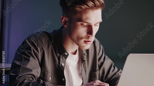 A young man types intently on a laptop, focusing on the task at hand in a dimly lit room. The man’s attention to the laptop reflects his dedication to completing important work.
