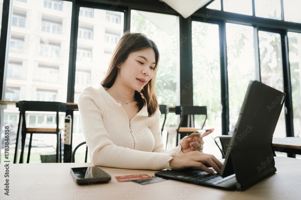 Obraz premium Young woman using a credit card to make online payments on her laptop in a modern cafe, representing digital lifestyle and e-commerce convenience.