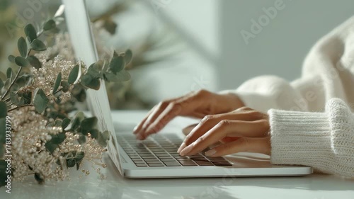 Closeup of female hands working on a laptop at a bright desk with eucalyptus leaves, evoking tranquility and focus. The scene highlights modern remote work and digital creativity in a serene setting