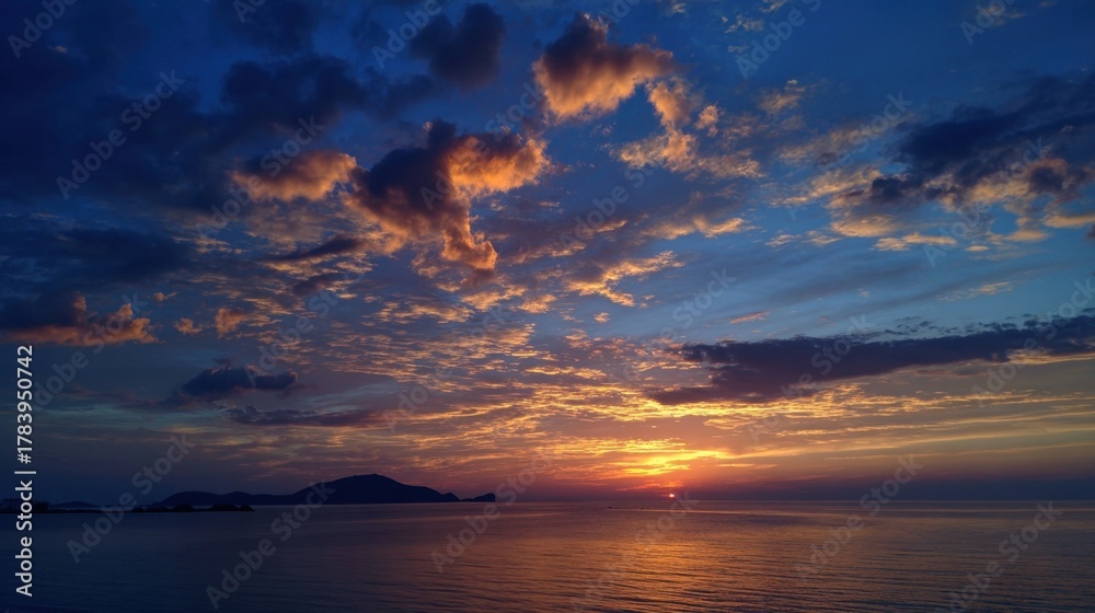 Naklejka premium A stunning sunset casts warm colors across the sky, reflecting on the calm ocean waters. Distant islands create a serene backdrop as day transitions to night.