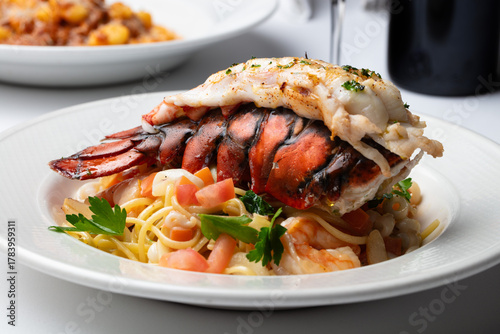 A plate of cooked lobster and pasta on a white clothed table with a red wine bottle and glass. 
