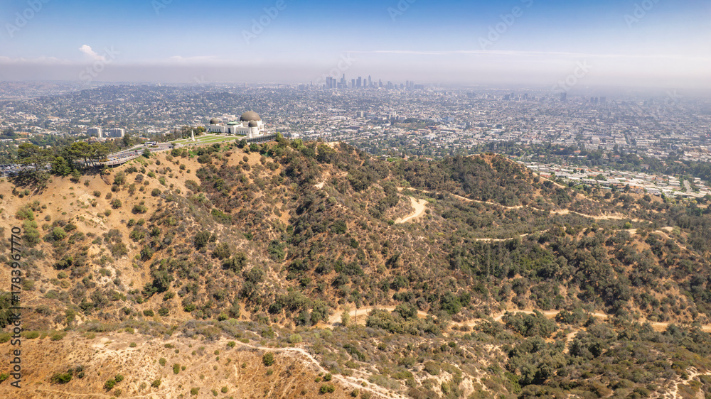 Obraz premium Drone shot of Griffith Observatory perched atop a hill with expansive Los Angeles city view. Dry vegetation, winding path, bright blue sky, urban sprawl, California landscape, iconic landmark