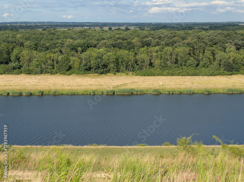 Wide calm river flanked by golden fields and dense green forest under blue sky, serene landscape.