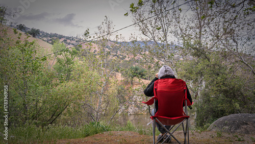 A lone backpacker sits in a folding chair overlooking a serene river valley. Moody landscape, reflection, contemplation, nature, peaceful moment.
