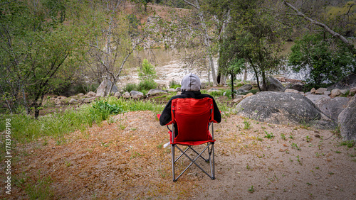 A lone backpacker sits in a folding chair overlooking a serene river valley. Moody landscape, reflection, contemplation, nature, peaceful moment.