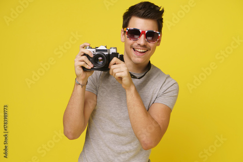 A young man in sunglasses smiles brightly as he holds up a vintage camera, capturing a moment in time