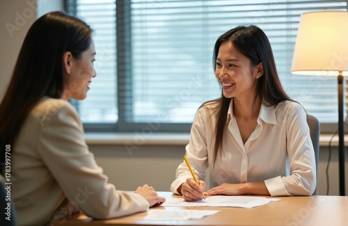 Two asian women sit at desk talk in office. Female manager interviews client. Employee writes with pencil on paper. Smiling women discuss project at workplace near window.
