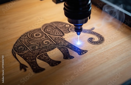 Laser engraver creates detailed elephant picture on wood. Tech tool burns image, with smoke curls. Concept includes industrial design, craft and manufacture with machinery on light brown wood surface.