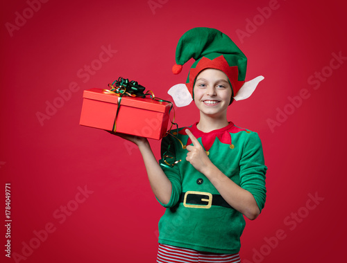 Merry Christmas concept. Smiling funny boy in Santa's elf costume holding Christmas gift in his hand. Shooting on red background.
