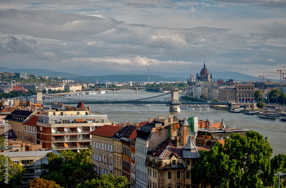 Naklejka premium Budapest, Hungary - July 28, 2025: Panoramic view of the Danube River flowing through central Budapest, with the Hungarian Parliament Building visible on the right and city architecture lining the riv