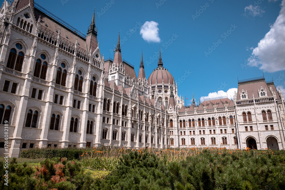 Obraz premium Budapest, Hungary - July 27, 2025: The Hungarian Parliament Building viewed from its garden side, showcasing its magnificent Neo-Gothic architecture with detailed spires and ornate stonework.