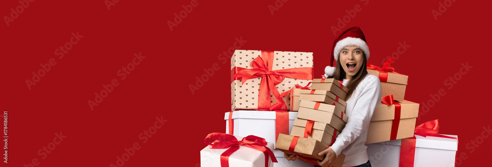 Obraz premium A cheerful woman wearing a Santa hat stands among colorful wrapped gifts, expressing joy and excitement. The background features a vibrant red color, adding to the festive atmosphere.