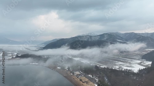 Fly over misty winter rice fields and snow-dusted lakes in Fukushima, with distant mountains standing majestically. This tranquil aerial view captures Japan’s serene countryside, meditation.