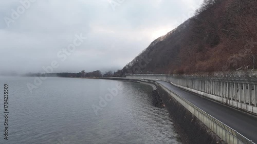 Aerial view of a lakeside road at Lake Inawashiro. Gradually rising and moving forward, the scene transitions from the road to misty fields, serene lake, and ethereal fog, perfect for meditation.