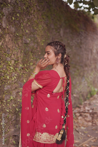 A young woman wearing a traditional red salwar kameez with golden embroidery stands outdoors near an old stone wall covered with natural moss