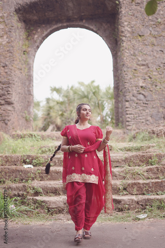A young woman wearing a traditional red salwar kameez with golden embroidery stands outdoors near an old stone wall covered with natural moss