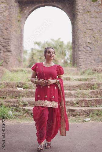 A young woman wearing a traditional red salwar kameez with golden embroidery stands outdoors near an old stone wall covered with natural moss