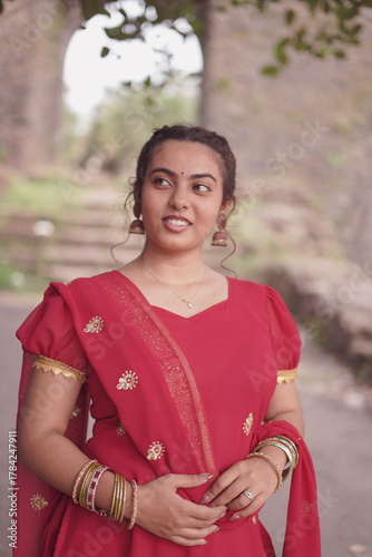 A young woman wearing a traditional red salwar kameez with golden embroidery stands outdoors near an old stone wall covered with natural moss