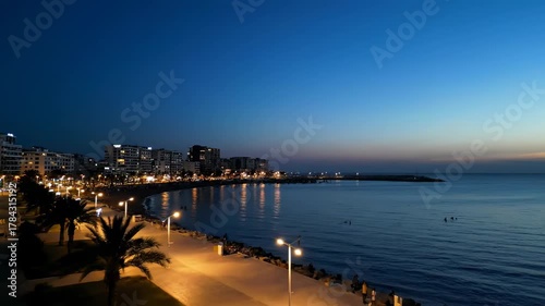 A coastal city at dusk features buildings along a shoreline, palm trees, streetlights, and a calm sea mirroring city lights. A serene night scene