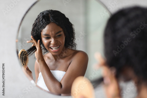 Photos Irritated black lady with bushy hair stands in front of a bathroom mirror, using a wooden brush