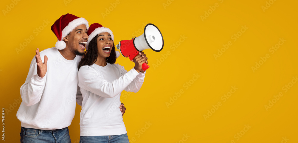 Obraz premium A cheerful couple wears Santa hats while excitedly holding a red megaphone. They are celebrating together against a vibrant yellow backdrop, showcasing a joyful holiday spirit.