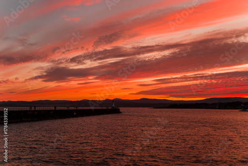 A vibrant sunset over the Pacific coast, the sky is saturated with vibrant colors, the horizon is marked by distant hills. The composition is serene and dramatic due to the contrast and natural beauty
