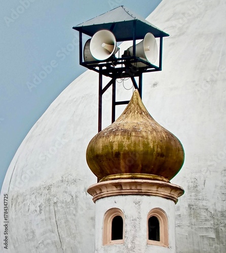 A mosque dome with a golden finial topped by loudspeakers against a pale sky, reflecting traditional Islamic architecture combined with modern elements for prayer calls and community gatherings.
