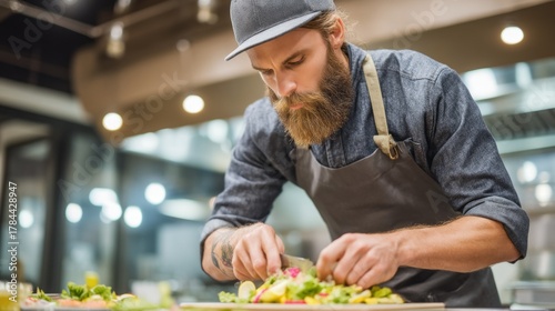 In a contemporary kitchen, a bearded chef in a stylish apron meticulously arranges a colorful salad. The warm evening light enhances his focused expression as he prepares a delightful dish