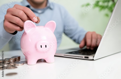 Fototapeta Naklejka Na Ścianę i Meble -  Man holding coin, putting it into a pink piggy bank while using a laptop for online banking or managing personal finance