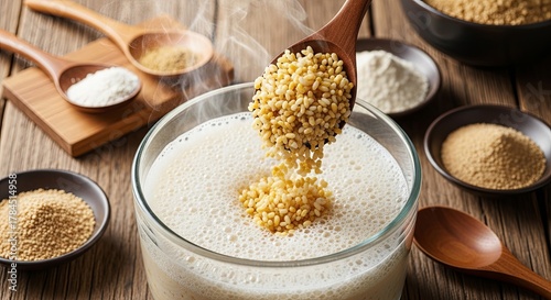 Fresh millet grains pouring from wooden spoon into glass bowl of foamy plant milk with various gluten free flour powders and ingredients on rustic wooden kitchen counter
