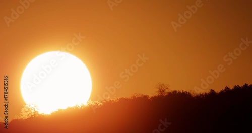 Sunrise time lapse, massive bright orange sun rising rapidly over a dark mountain forest silhouette catching the vivid morning golden hour 4K