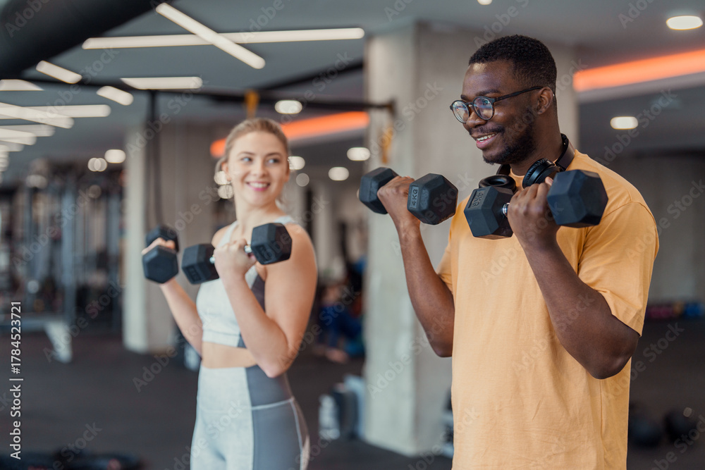 Fototapeta premium Indoor gym scene with smiling diverse young adults exercising with dumbbells in casual athletic wear