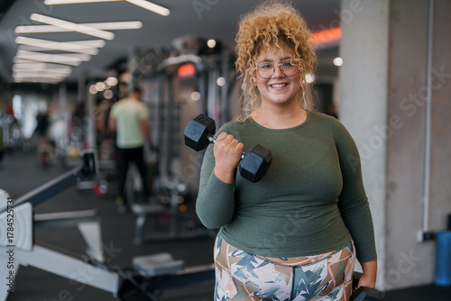 Smiling plus size woman in green long sleeve top holding dumbbell exercises indoors at gym