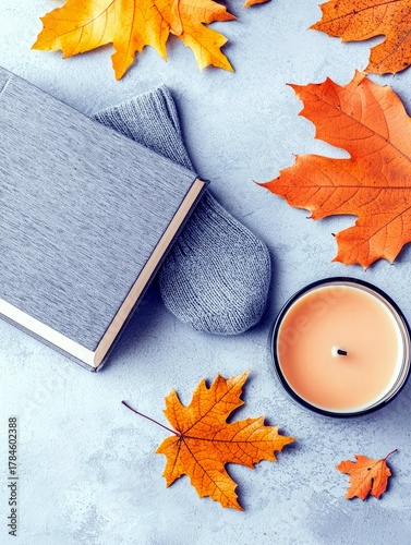 A flat lay composition of a book, gray socks, a lit candle, and vibrant orange and yellow autumn leaves on a textured gray surface.
