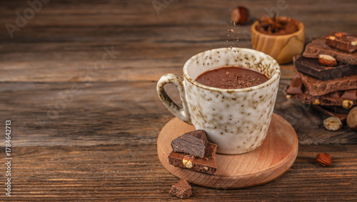 Cup of hot chocolate on wooden table with falling cocoa powder, pieces of dark and milk chocolate with hazelnuts, cozy rustic background with copy space for warm drink concept