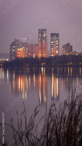 Evening view of Obolon Embankment in Kyiv on a cloudy day. City lights and tall buildings reflect in the calm Dnipro River, creating a peaceful and atmospheric urban scene.
