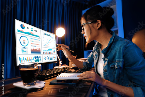 Smiling young beautiful asian creative looking on pc of profit of sales target report and writing on notebook for memo with planning invest new strategy market project at neon light night. Stratagem.